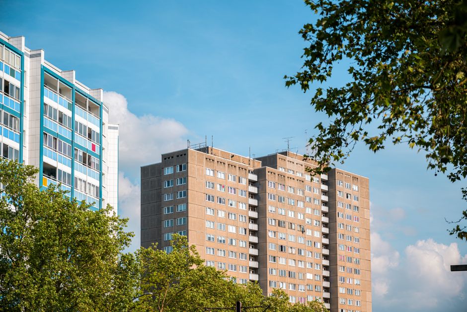 A shot of two multi-storey residential buildings in an urban area with clear blue sky and some scattered clouds. The building on the left features modern architecture with a white and blue facade, large glass windows, and balconies, while the building on the right is a taller, beige-colored block with numerous small windows and external staircases. In the foreground, green trees partially obscure the lower parts of the buildings. The scene captures a typical cityscape that may be involved in house removals or home relocation services. This setting could represent part of a process where furniture and packing materials are transported from the apartment buildings, with Elephant and Castle Removals providing professional moving and loading services in the area.