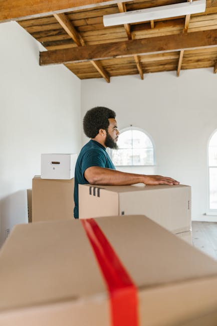 A man with dark skin and curly black hair, dressed in a navy blue t-shirt, stands inside a bright, partially constructed or renovated room with a high wooden ceiling and exposed beams. He is leaning against a stack of large cardboard moving boxes, some of which are sealed with red packing tape, suggesting he is involved in packing or preparing for a home relocation. In the background, two arched windows allow natural light to enter, illuminating the space. Additional cardboard boxes are visible behind him, indicating an ongoing packing process in a house undergoing furniture transport or moving preparations. The scene captures a moment of loading or packing during a professional house removal, with the focus on precise handling and organization essential for a smooth relocation process, as managed by Elephant and Castle Removals.