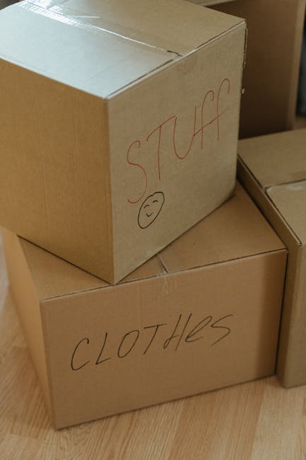 Close-up image of two light brown cardboard boxes used in home relocation, stacked on a wooden floor. The top box is labeled 'STUFF' in red marker with a small, simple smiley face drawn below the text. The lower box is labeled 'CLOTHES' also in red marker. The boxes are sealed with packing tape and are part of the packing process during furniture transport and packing and moving activities. In the background, additional boxes are partially visible, indicating an organized packing setup for a house remova. The scene suggests a careful staging of the moving process, with clear labels to facilitate sorting and unloading, supporting professional removal services like Elephant and Castle Removals operating in the SE1 area.