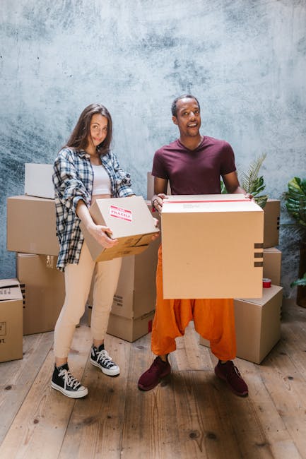 Two individuals inside a residential room engaged in a home relocation process, carrying cardboard boxes filled with household items. The woman on the left is holding a medium-sized box labeled 'Fragile' and wearing casual clothing including a plaid shirt, beige pants, and sneakers. The man on the right is holding a larger box and is dressed in a maroon t-shirt, orange trousers, and brown shoes, smiling while preparing for furniture transport. Surrounding them are additional packed cardboard boxes of various sizes stacked on the wooden floor, some sealed with packing tape. The background features a textured blue-grey wall, and to the right of the man, a potted plant adds a touch of greenery to the scene. The environment suggests an organized packing and moving process as part of a professional removals service by Elephant and Castle Removals, focusing on careful packing and efficient loading for a house move in the area, with the action taking place indoors in natural lighting.