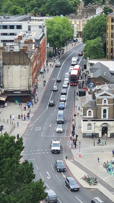 An aerial view of a busy urban street lined with mixed-use buildings, including red brick and modern white facades, with pedestrians walking along the sidewalks and crossing at designated crosswalks. Several cars and a double-decker bus are traveling along the road, which features clearly marked lanes and parking areas. On the right side of the image, a small building with a gabled roof is visible next to a bicycle parking area, while large green trees provide shade and greenery along the street. This scene captures a typical city environment suitable for home relocation and furniture transport logistics, with vehicles and pedestrians supporting the movement of household items during a move with Elephant and Castle Removals.